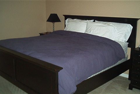 1985 Ford LTD Crown Victoria - A bedroom scene features a large bed with a dark wood frame and a purple comforter, set against a neutral-colored wall.