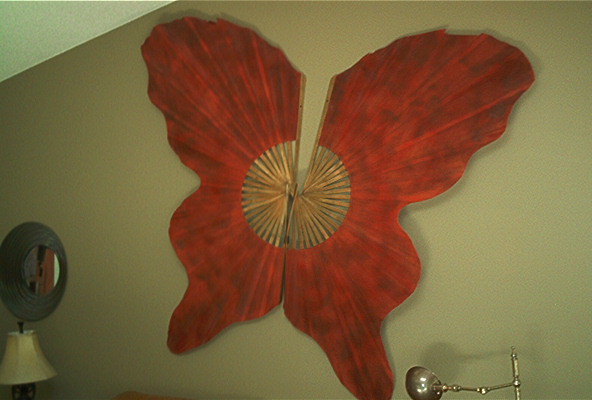 Red butterfly-shaped wall art with a wooden fan center, mounted on a light green wall. A lamp and mirror are visible to the left