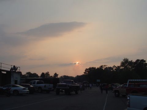 1979 Ford Bronco - Sunset over a gravel lot at a truck event, with a lifted square-body pickup on large tires silhouetted in the center and a white extended-c…
