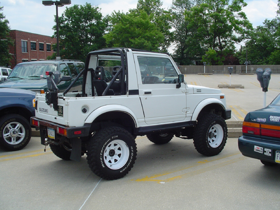 White Suzuki Samurai with lifted suspension and off-road tires parked in a lot next to a blue SUV and a dark sedan