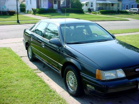 1989 Ford Taurus - A dark sedan is parked on the grass and curb in front of a house on a sunny day.
