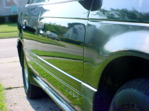 1989 Ford Taurus - Side profile of a dark green sedan with chrome trim, reflecting a suburban street scene. Watermark reads "SUPERMOTORS".