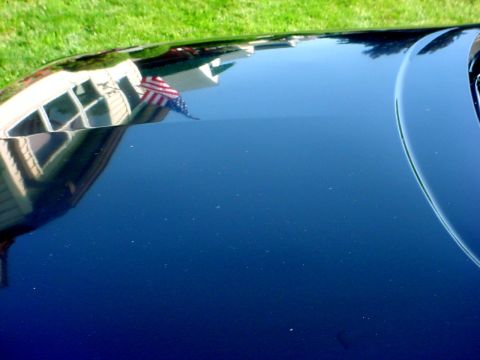 1989 Ford Taurus - The glossy surface of a car hood reflects a house and an American flag under a bright sky.