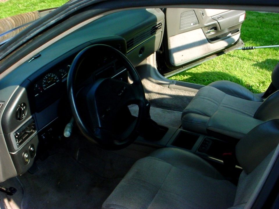 Interior shot of a car showing the dashboard, steering wheel, and front seats. The door is open, revealing grass outside