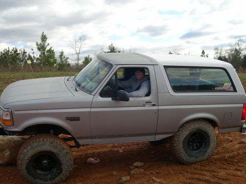 1995 Ford Bronco - A man drives a silver Ford Bronco with large tires through a dirt landscape.
