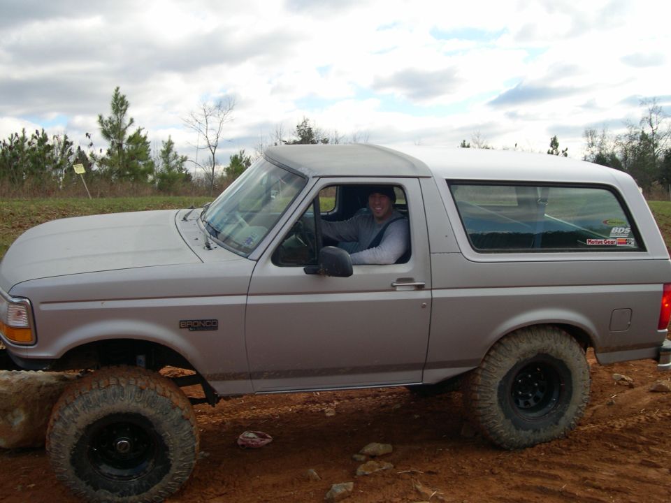 Silver Ford Bronco with off-road tires on a dirt path, driver visible