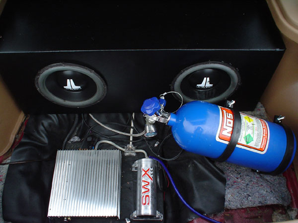 Trunk with two JL Audio subs in a black box, silver amp, SWX capacitor, and blue NOS bottle.