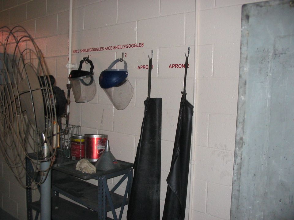 Face shields, goggles, and aprons hang on a wall above a shelf with cans and a funnel. A large fan is on the left