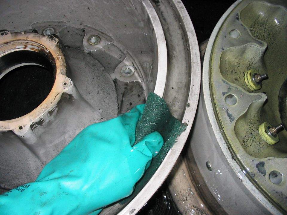 Gloved hand scrubs a metal brake drum assembly. Another drum is visible to the right