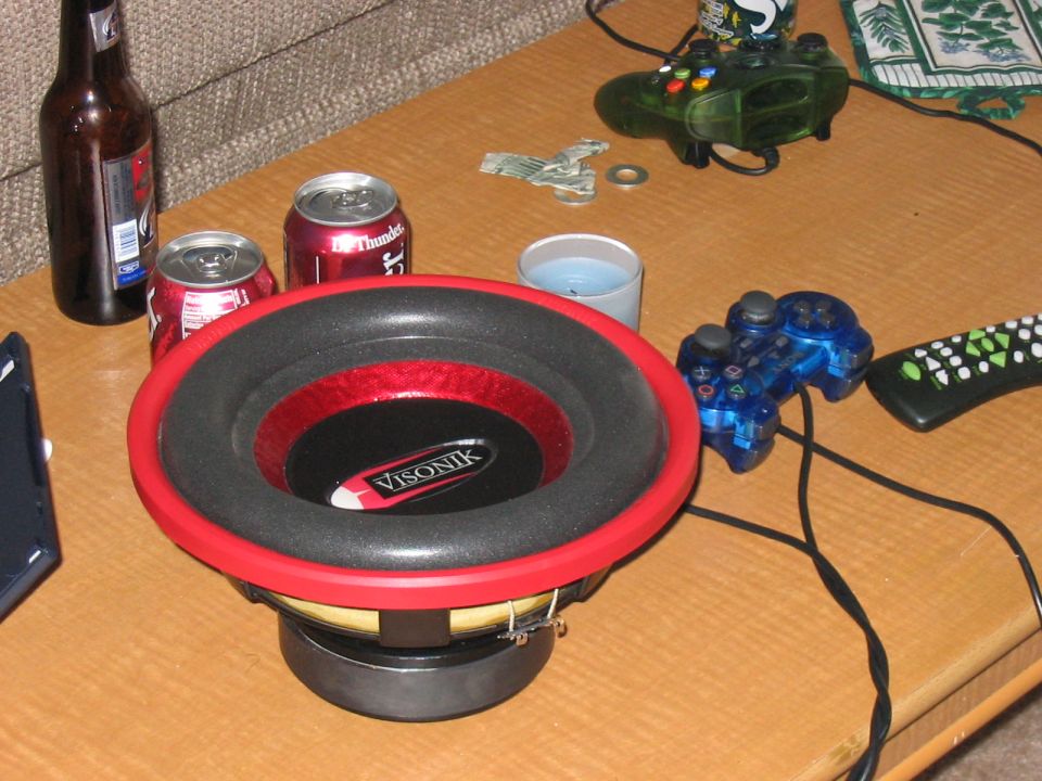 Car subwoofer with red trim, two cans of soda, a beer bottle, a candle, and video game controllers on a table