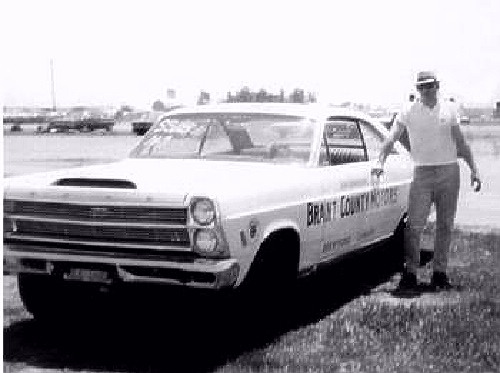 Vintage B&W photo of white 1960s Ford coupe with man leaning on it at a track