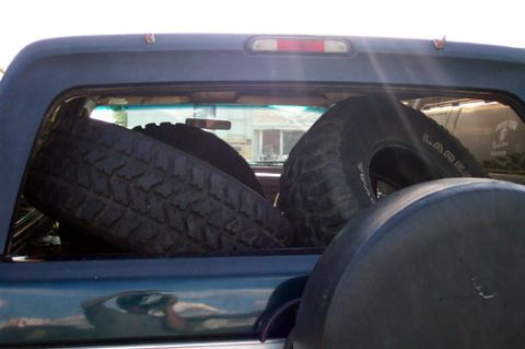 1995 Ford Bronco - Three aggressive mud-terrain tires loaded loose in the bed of a dark blue/teal pickup, shot from behind the cab with the rear window and th…