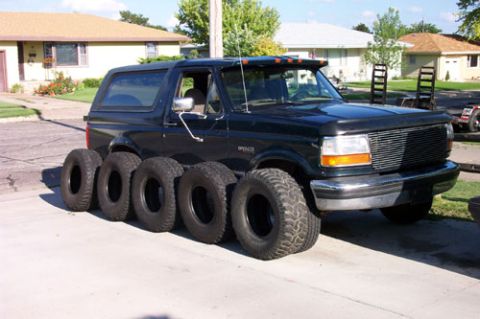 1995 Ford Bronco - Black two-door full-size SUV parked in a residential driveway with a row of five spare off-road tires and wheels lined up on the pavement i…