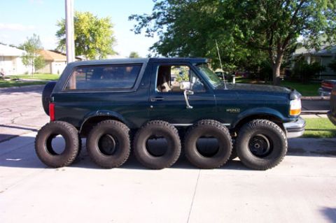 1995 Ford Bronco - Side profile of a dark green Ford Bronco built as a novelty rig, stretched onto five axles with ten off-road tires lined up along the pavem…