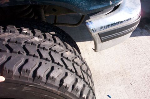 1995 Ford Bronco - A tall all-terrain/mud-terrain tire leaned tread-first against the ground next to the chrome rear step bumper of a dark green truck, showin…
