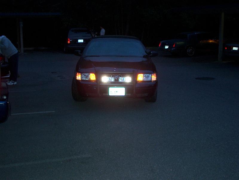 Front view of a red sedan with headlights on. Other cars are parked in the background