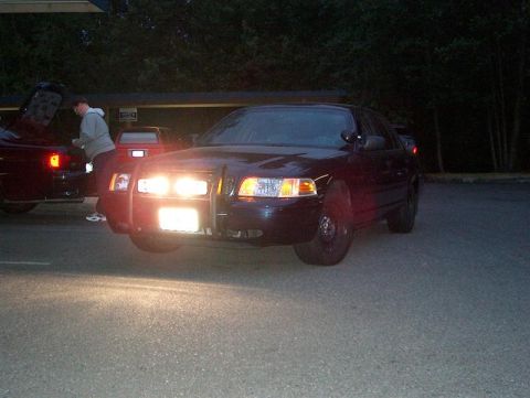 2001 Ford Crown Victoria - A dark sedan with a push bumper is parked at night, with another vehicle visible to the left.