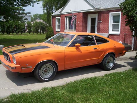 1978 Ford Mustang II - This orange coupe with racing stripes is parked in front of a brick house.