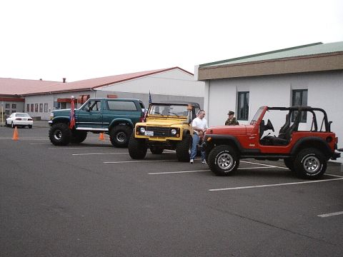 1996 Ford Bronco - Three modified off-road vehicles are parked in a lot, with two people standing near the yellow Jeep.