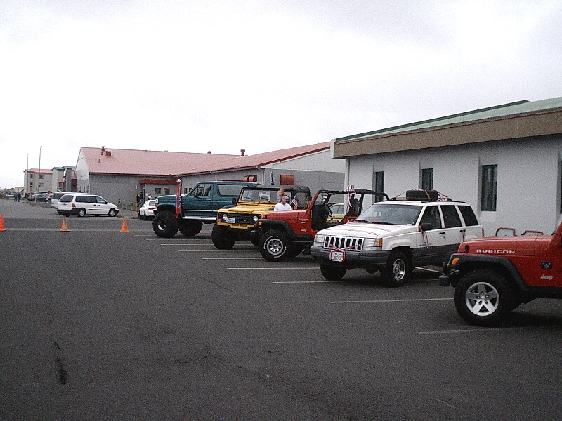 Row of Jeeps and a lifted teal pickup parked in a lot by warehouse buildings.