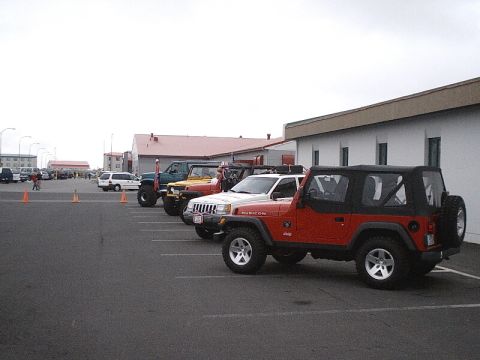 1996 Ford Bronco - Several Jeep Wranglers are parked in a lot in front of buildings under a cloudy sky.