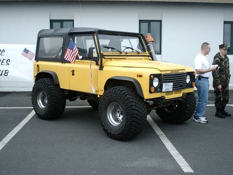 1996 Ford Bronco - A yellow Land Rover Defender is parked on asphalt with two men standing next to it.