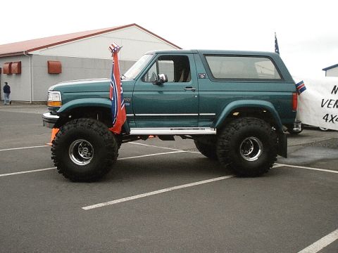 1996 Ford Bronco - A modified green 1996 Ford Bronco is parked on asphalt, oversized tires and a flag mounted on the front fender.