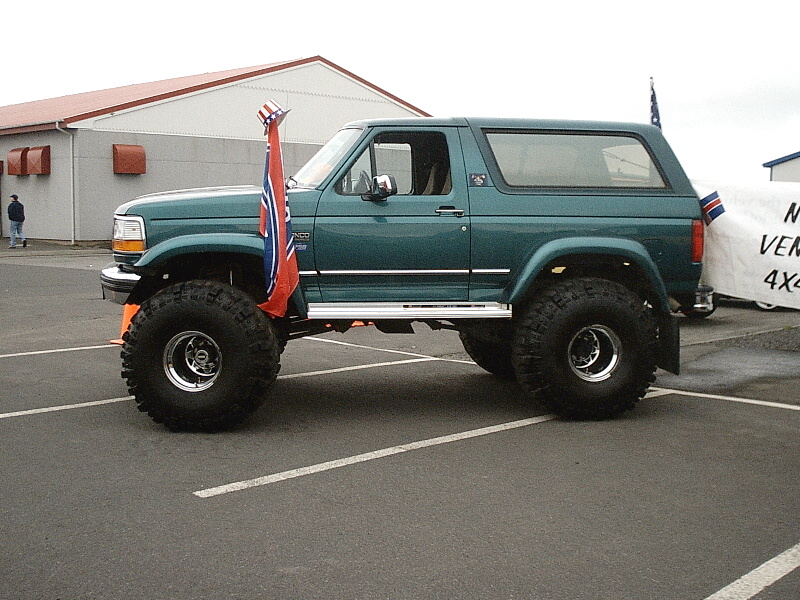 Green lifted two-door Ford Bronco on large off-road tires, side view in a parking lot.