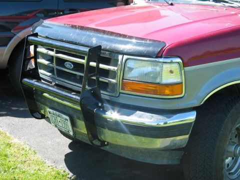 1995 Ford Bronco - Close-up 3/4 front view of a red and tan Ford SUV with a black tubular brush guard/grille guard mounted over the front end, showing the For…