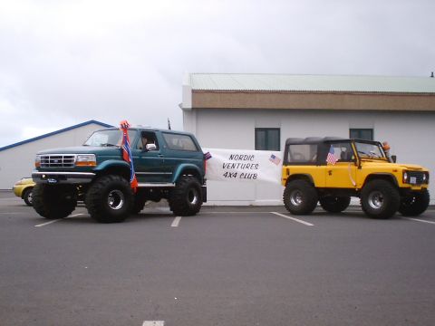 1996 Ford Bronco - Two off-road vehicles, a teal SUV and a yellow Defender, are parked outside a building with a banner for Nordic Ventures 4x4 Club.