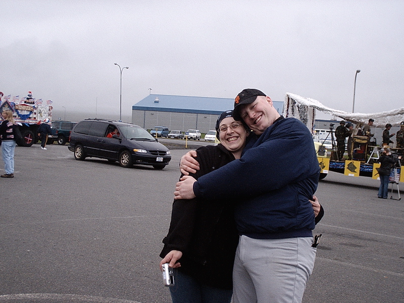 Two smiling people hugging in a parking lot with a minivan and parade floats behind them