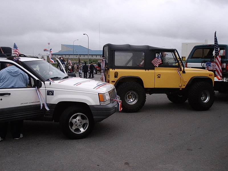 Yellow soft-top 4x4 parked between a white Jeep and flag-decorated vehicles at an outdoor event