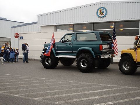 1996 Ford Bronco - A modified green Ford Bronco with oversized tires and flags is parked next to a yellow vehicle.