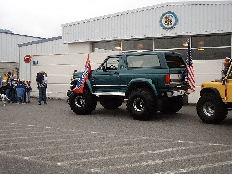 Lifted teal two-door Ford Bronco on big off-road tires, rear 3-quarter view in a parking lot with a flag.