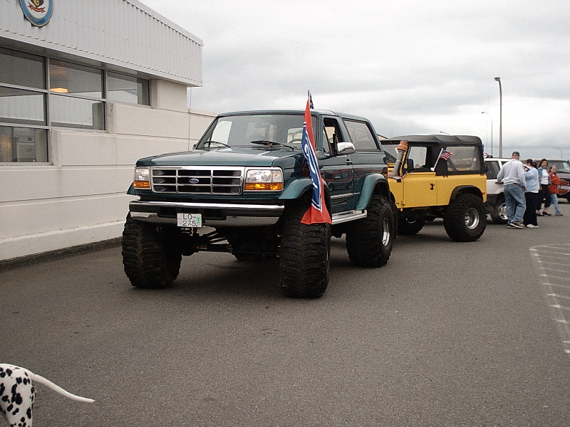 Dark green lifted Ford Bronco on big tires parked in a lot with a flag and other SUVs behind.