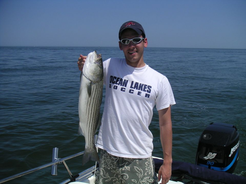 Man in white Ocean Lakes Soccer shirt and sunglasses holds striped bass on a boat at sea