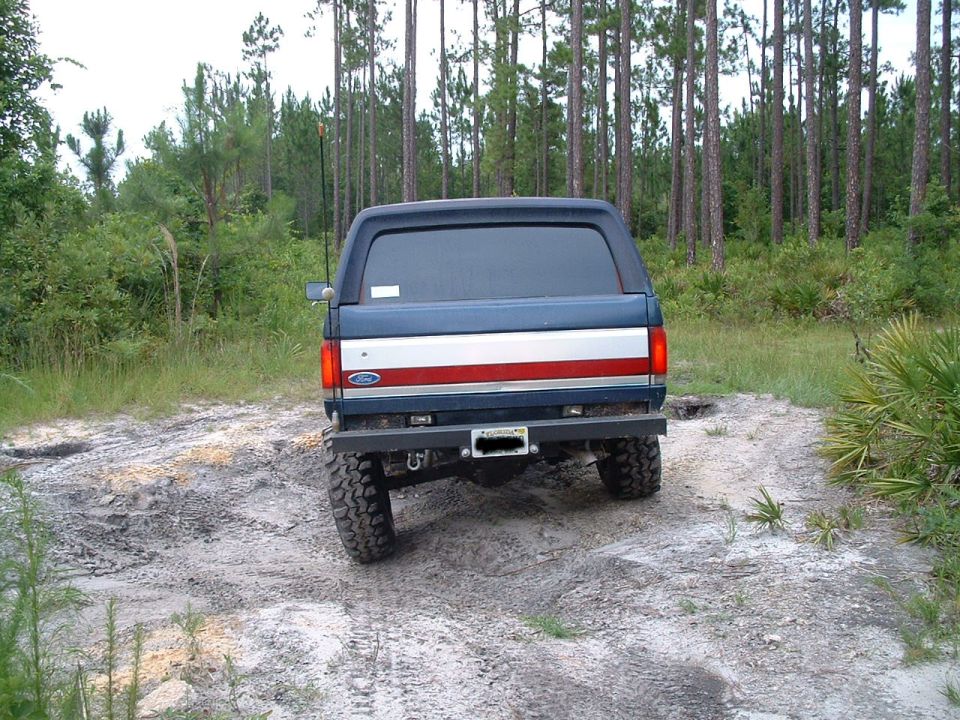 Ford Bronco on dirt road, rear view. Blue and red stripe on tailgate. Aftermarket bumper