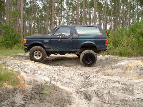 1995 Ford Bronco - A lifted Ford Bronco navigates a dirt path surrounded by trees.