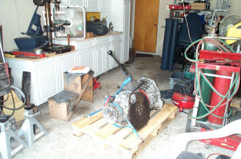 1995 Ford Bronco - A vehicle transmission with attached shifter and flywheel/torque converter sits strapped to a wooden pallet on a garage floor. The surround…