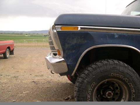 1978 Ford Bronco - A blue pickup truck with off-road tires sits in a dusty lot, with a red truck in the distance under a cloudy sky.