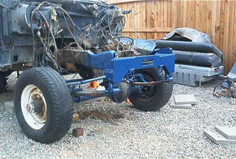 1986 Ford Bronco - A partially disassembled vehicle chassis with a freshly painted blue front frame section and axle, mounted on white steel wheels with worn…