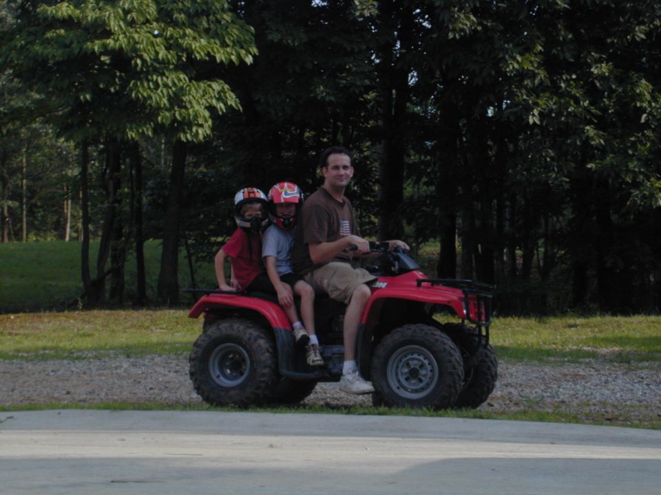 Dad driving a red ATV with two kids in helmets in front of trees