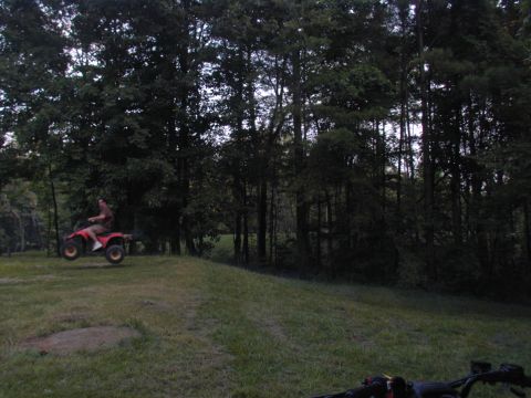 1988 Ford Bronco - A person rides a red ATV as it becomes airborne over a grassy hill with trees in the background.