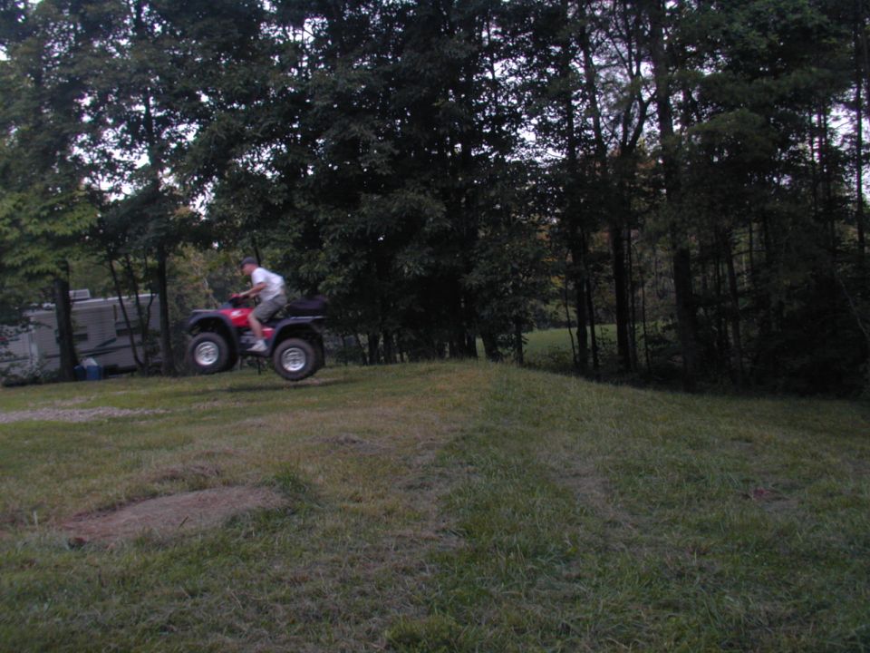 Red ATV airborne over a grassy mound with trees and a trailer in the background