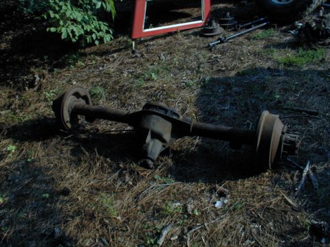 1988 Ford Bronco - A rusty vehicle axle assembly rests on the ground amidst debris and foliage.