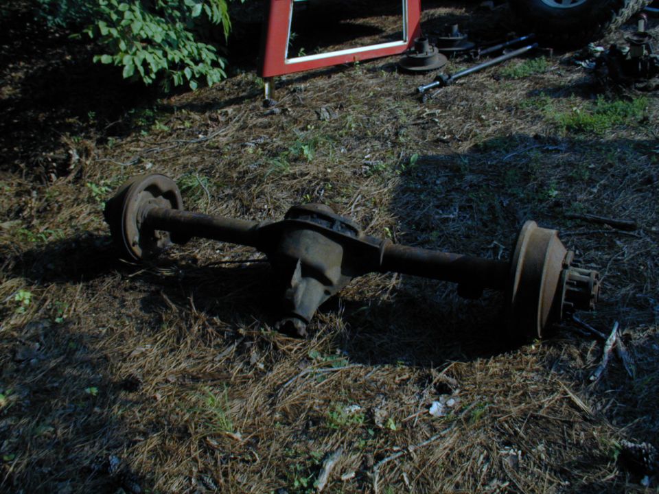 Old axle assembly lying on the ground with some red metal and tires in the background
