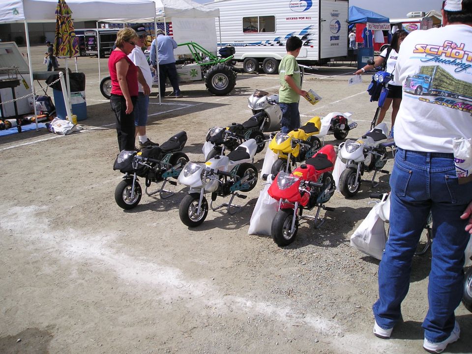 Several mini motorcycles lined up on dirt ground, people standing around them