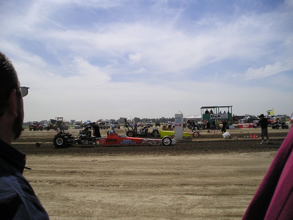 Dragsters lined up on a dirt track with spectators and support vehicles