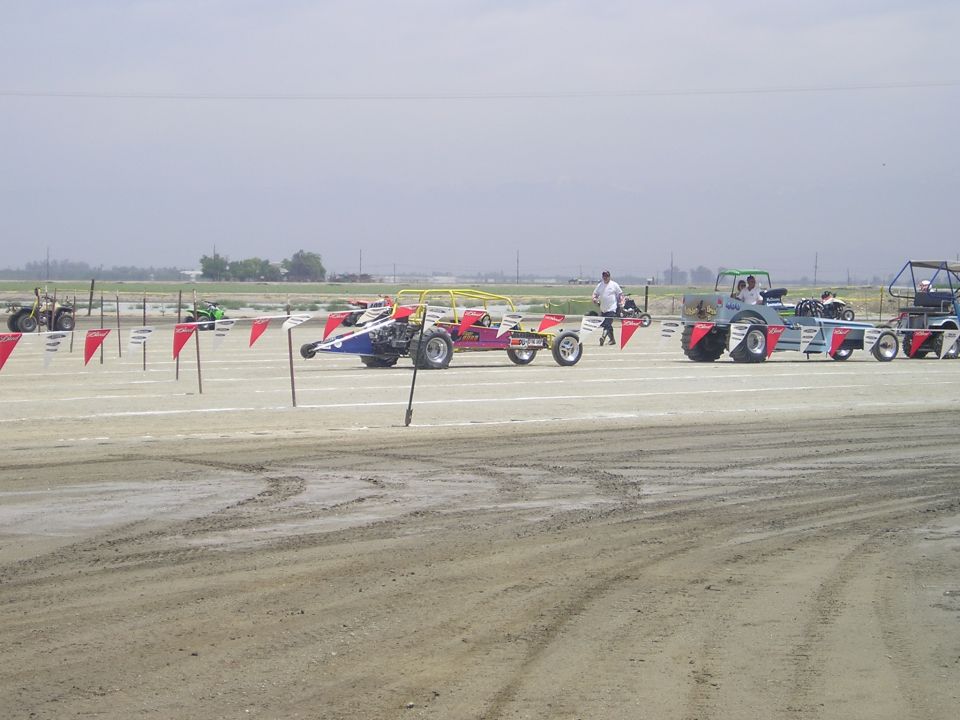 Multiple drag racing vehicles lined up on a dirt track under a cloudy sky