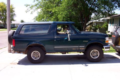 1995 Ford Bronco - Driver-side profile of a dark green Ford Bronco full-size SUV with matching removable hardtop, parked at the curb in a leafy residential ne…